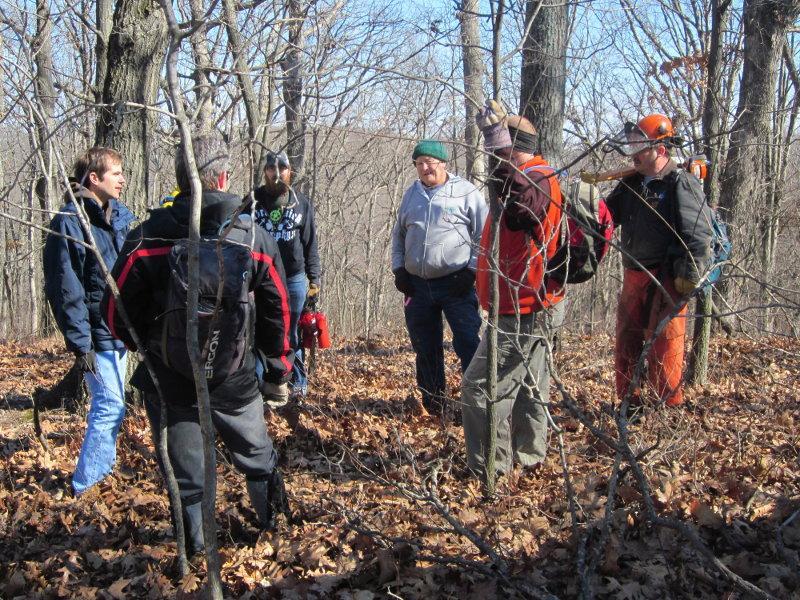Group of six people standing in a wooded area during fall or winter, surrounded by bare trees and fallen leaves. They appear to be engaged in a discussion or meeting, with some wearing outdoor gear and gloves. One person holds a tool, and another has a backpack. The atmosphere suggests a focus on outdoor activities or conservation. Greensfelder Park mountain bike trail.