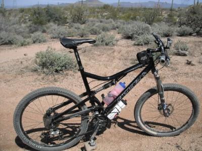 Santa Cruz Superlight: A black mountain bike with a water bottle attached, parked on a dirt trail surrounded by desert vegetation and shrubs. In the background, there are distant hills and a clear blue sky.