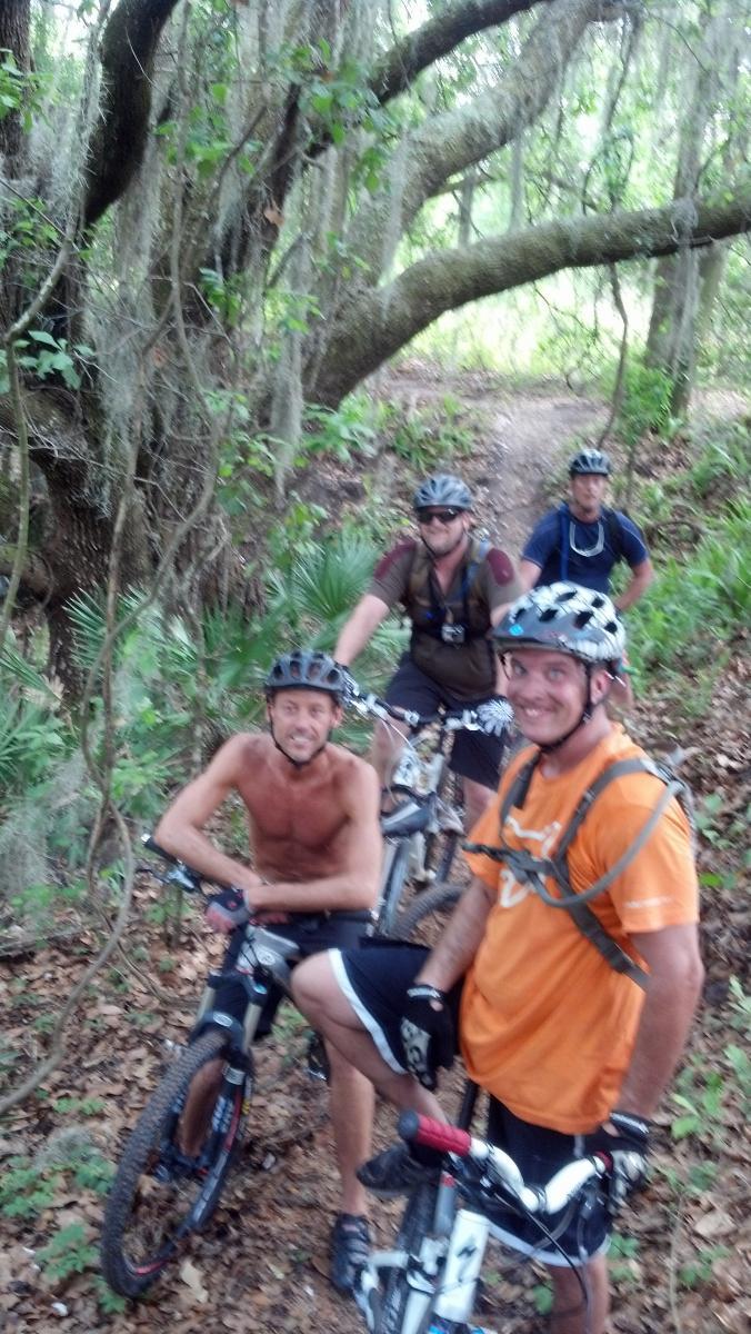 Four mountain bikers pose on a wooded trail, surrounded by lush greenery and large trees. One rider, shirtless, smiles at the camera, while another wears sunglasses and a helmet. The group appears relaxed and friendly, with their bikes nearby, ready for their adventure. Balm Boyette Scrub Preserve mountain bike trail.