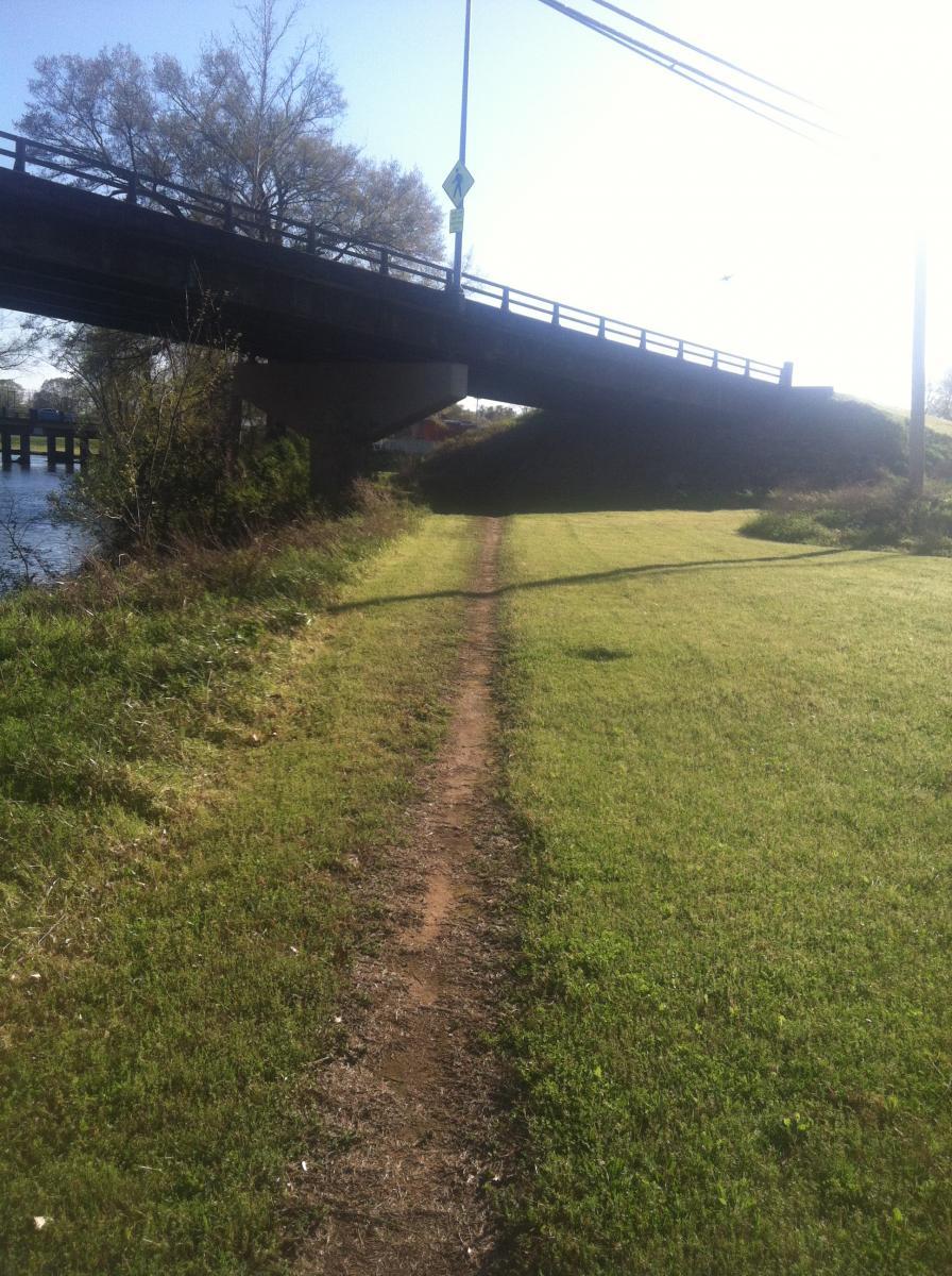 A grassy path leading through a park area, located under a bridge. The scene includes a river visible to one side and clear blue skies above, with a few trees lining the path. The pathway is slightly worn, indicating foot traffic, and there are road signs hanging from the bridge above. Spadra Creek Nature Trail mountain bike trail.