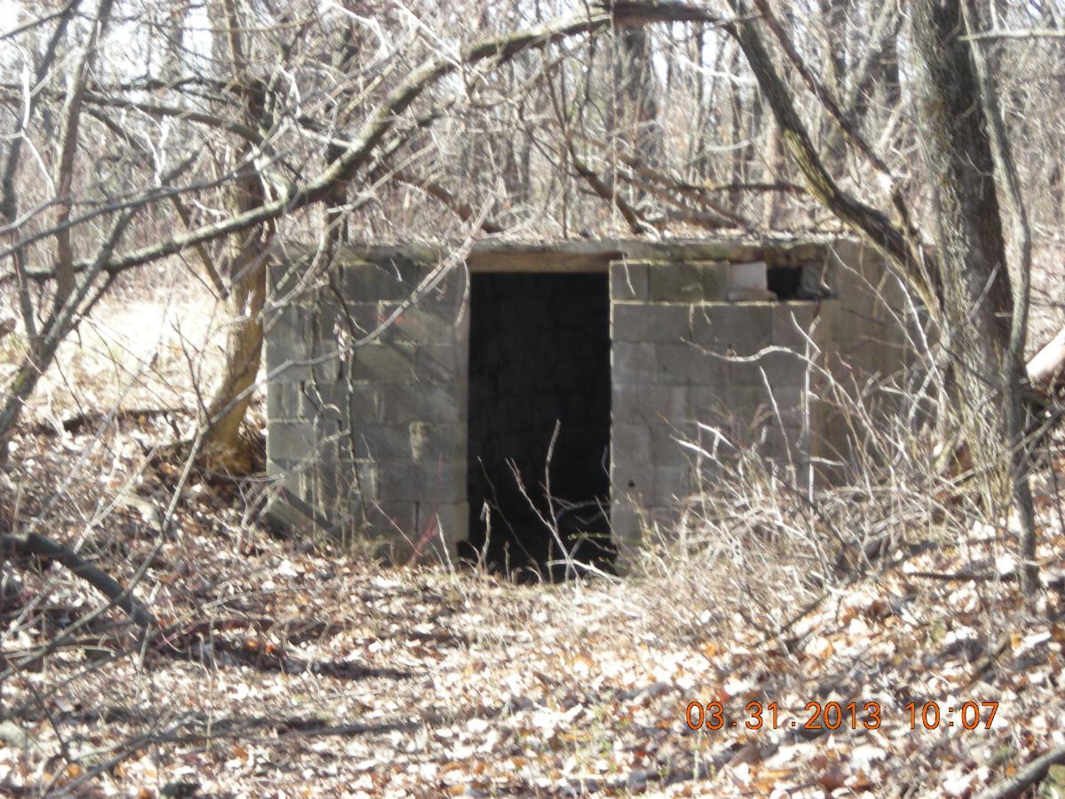 A small, dilapidated concrete structure partially hidden by overgrown vegetation in a wooded area. The entrance is partially visible, and the surrounding ground is covered with dried leaves and branches. Melvern Riverfront Trails mountain bike trail.