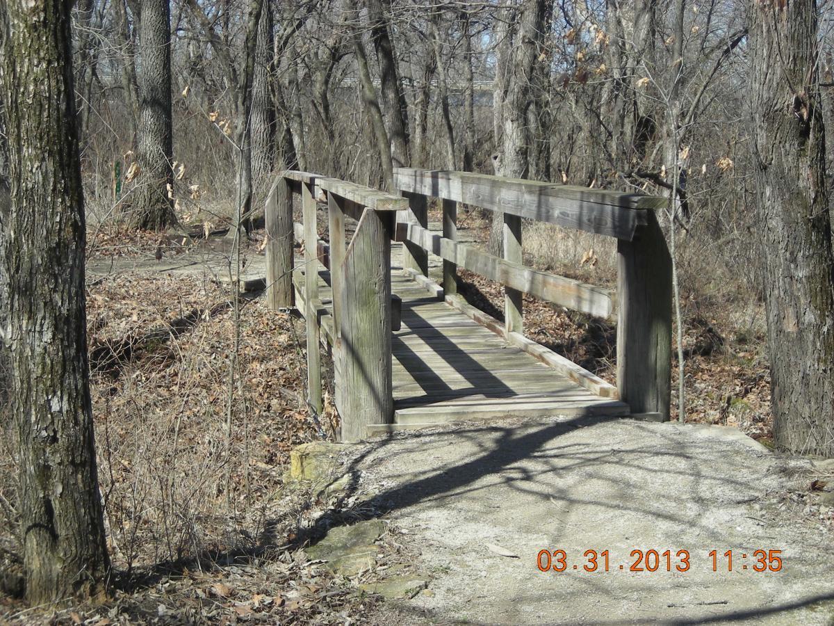 A wooden footbridge spans a small path through a wooded area, surrounded by bare trees and dried leaves on the ground. The bridge features a simple design with railing on both sides, inviting passage across the natural setting. Melvern Riverfront Trails mountain bike trail.