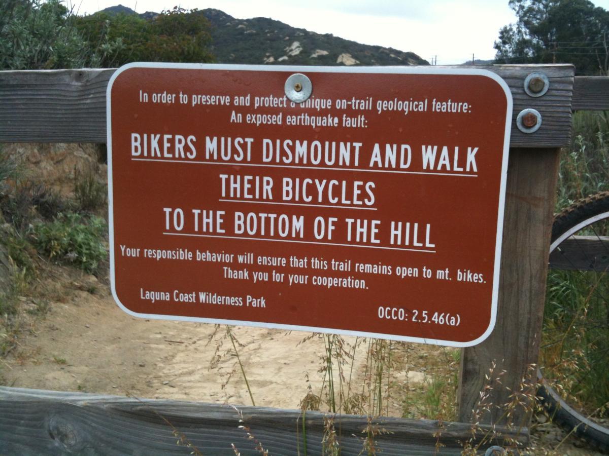 Sign at Laguna Coast Wilderness Park instructing bikers to dismount and walk their bicycles to the bottom of the hill to protect an exposed earthquake fault. Stagecoach Trail (South) mountain bike trail.
