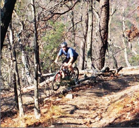 A mountain biker navigating a dirt trail through a wooded area, surrounded by bare trees. The cyclist is wearing a helmet and is in a dynamic riding position as they maneuver over a fallen log on the trail. Douthat State Park mountain bike trail.