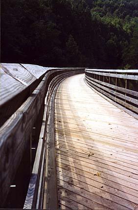 A wooden walkway curves gently through a natural landscape, surrounded by lush green trees. The path is lined with wooden railings and has a smooth, sunlit surface, inviting outdoor exploration. Greenbrier River Trail mountain bike trail.