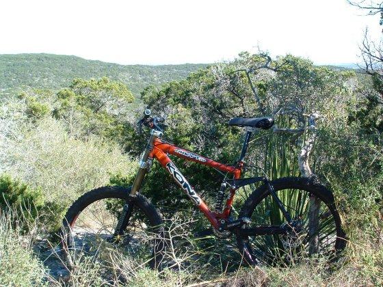 A mountain bike with an orange and black frame is leaning against a rock amidst a lush landscape of greenery and low bushes, with rolling hills visible in the background under a clear blue sky. Government Canyon mountain bike trail.
