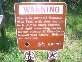 A warning sign for an advanced mountain bike trail, featuring text that describes the trail's characteristics, including short switchback climbs, steep descents, and tight traversing curves. The sign advises against attempting the ride if it exceeds the rider's ability and states that helmets are required. It also indicates the trail distance of 5.67 miles, accompanied by bicycle and helmet icons. York River State Park mountain bike trail.