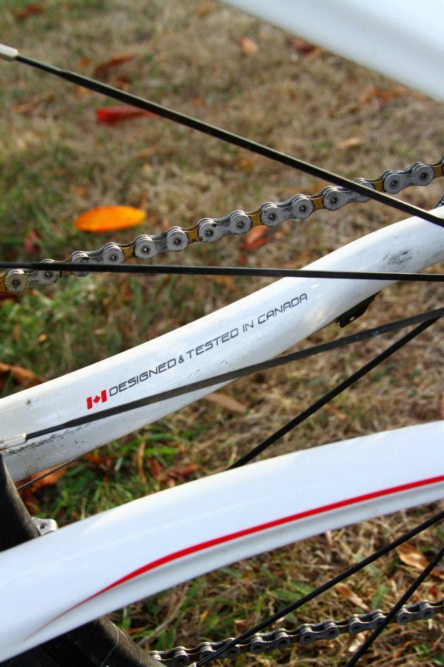 Rocky Mountain Vertex 50: Close-up image of a bicycle frame showing the text "DESIGNED & TESTED IN CANADA" along with a small Canadian flag, with a chain and wheel partially visible in the background. The scene features a grassy area with fallen leaves.