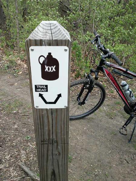 A wooden trail marker with a directional arrow and an illustration of a water jug, labeled "XXX," indicating the trail head. A mountain bike is parked next to the marker, surrounded by greenery and a dirt path. Creve Couer Park mountain bike trail.