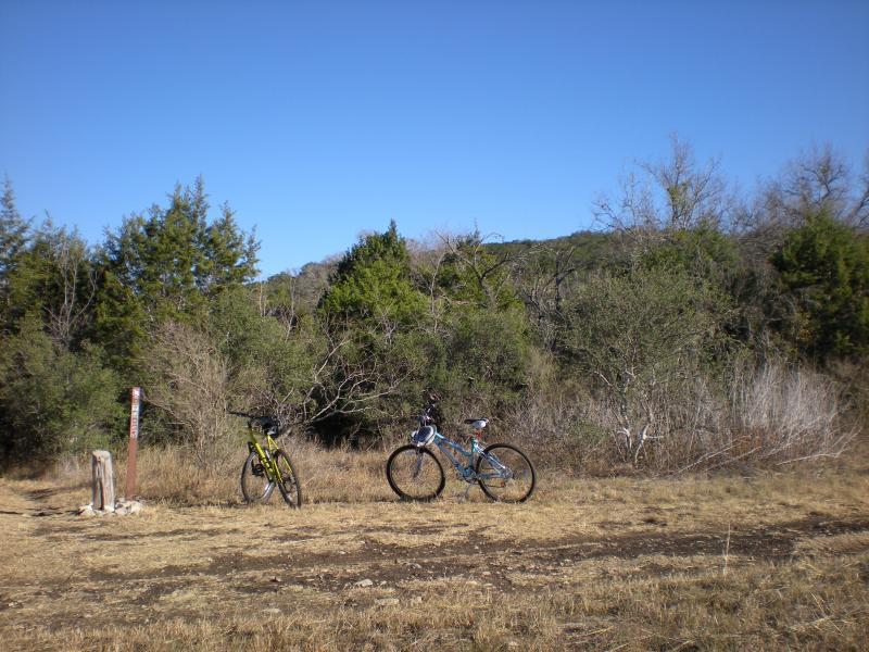 Two mountain bikes are parked on a dirt trail surrounded by sparse shrubs and trees under a clear blue sky. A wooden trail marker stands nearby. Government Canyon mountain bike trail.