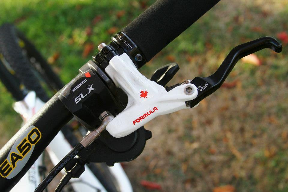Rocky Mountain Vertex 50: Close-up image of a bicycle handlebar showcasing a Shimano SLX gear shifter and a Formula brake lever. The handlebar features a black grip and a sleek design, with a white brake lever marked with a red maple leaf, set against a blurred background of grass and foliage.
