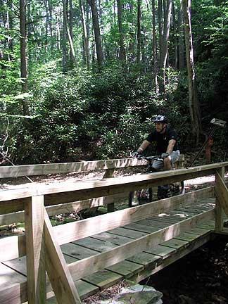 A mountain biker wearing a helmet rides across a wooden bridge in a forested area, surrounded by lush green trees and bushes. The scene showcases a peaceful outdoor environment ideal for biking and exploring nature. Coopers Rock mountain bike trail.