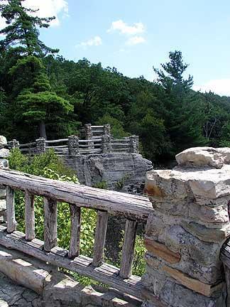 A rocky overlook with a wooden railing, surrounded by lush green trees under a blue sky with scattered clouds. Coopers Rock mountain bike trail.