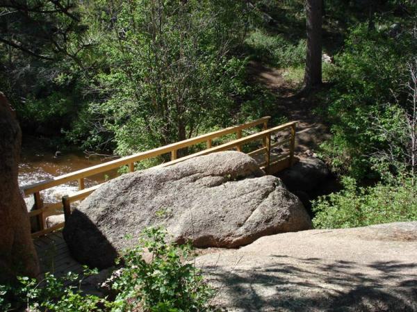 A peaceful natural scene featuring a wooden bridge crossing a small stream, surrounded by lush greenery and large rocks. The sunlight filters through the trees, illuminating the area, creating a tranquil outdoor setting. Curt Gowdy State Park mountain bike trail.