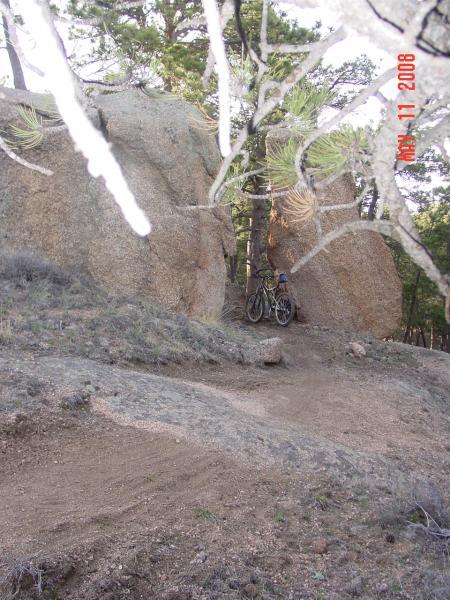 A mountain biking trail surrounded by large rocks and trees, with two bicycles leaning against a tree on the left. The terrain is uneven and dusty, showcasing a natural outdoor setting. Curt Gowdy State Park mountain bike trail.