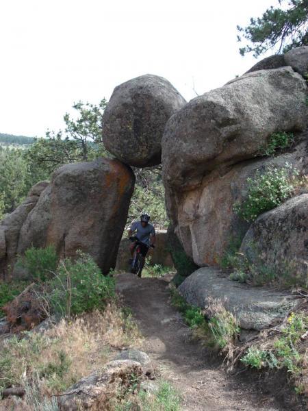 A mountain biker navigates through a narrow passage formed by large boulders on a dirt trail surrounded by greenery and trees. Curt Gowdy State Park mountain bike trail.