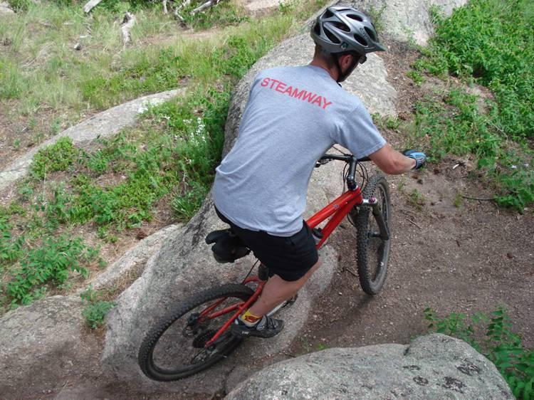 A person riding a mountain bike along a rocky trail surrounded by greenery. The rider is wearing a helmet and a grey shirt with "STEAMWAY" written on the back. Curt Gowdy State Park mountain bike trail.