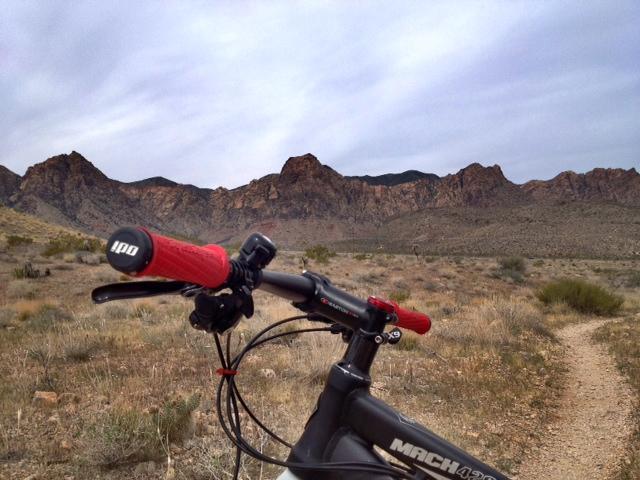 A close-up view of a mountain bike's handlebars with red grips, overlooking a sandy trail leading into a rocky mountain range under a cloudy sky. The landscape includes sparse vegetation, highlighting a rugged outdoor environment. Cottonwood Valley North mountain bike trail.