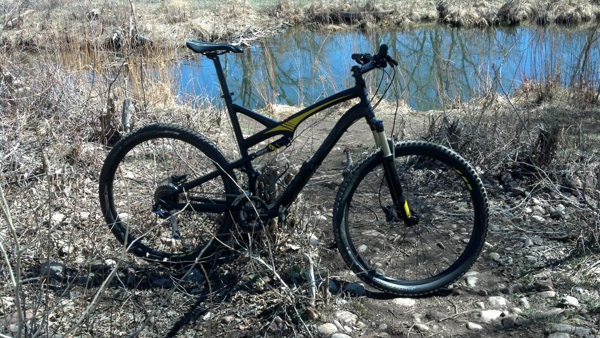 Specialized Camber 29: A black mountain bike with yellow accents is parked on a dirt trail surrounded by sparse vegetation and small rocks, near a calm body of water reflecting trees in the background.