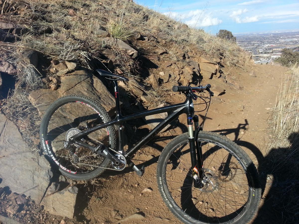 A black mountain bike resting on a rocky trail, surrounded by sparse vegetation and dirt. The background shows a panoramic view of a cityscape under a partly cloudy sky. Apex Park mountain bike trail.