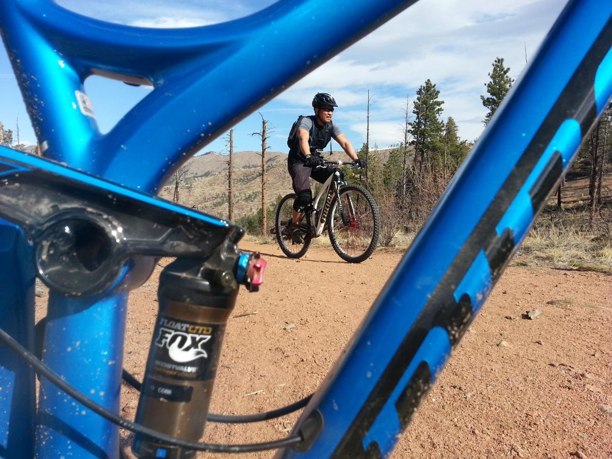A mountain biker riding on a dirt trail, viewed through the frame of a blue bicycle. The background features trees and a rocky landscape under a clear sky. The cyclist is wearing a helmet and is focused on the trail ahead. Buffalo Creek mountain bike trail.