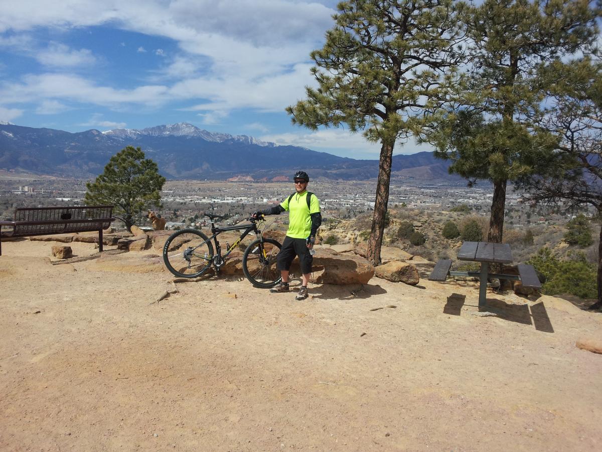 GT Karakoram: A person wearing a bright yellow safety vest stands next to a mountain bicycle on a rocky path overlooking a valley. Trees are visible nearby, and the backdrop features distant mountains and a blue sky with scattered clouds. A picnic table can be seen in the foreground, along with a bench in the background.