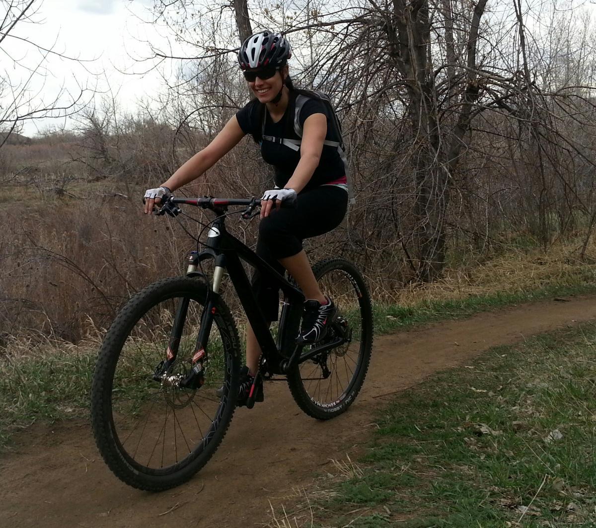 A person riding a mountain bike on a dirt trail surrounded by trees and tall grass, wearing a helmet and sunglasses, and smiling. The scene is set outdoors on a cloudy day. Bear Creek Lake Park mountain bike trail.