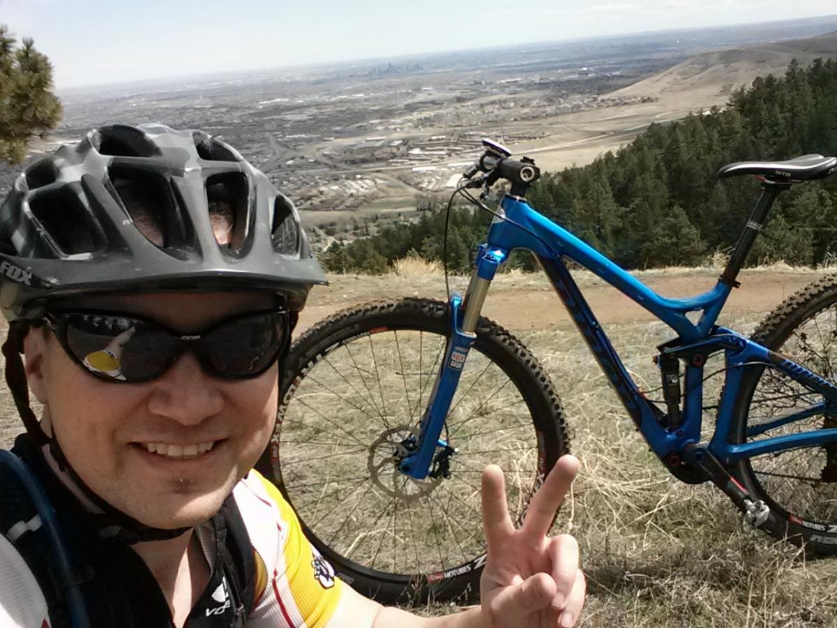 A person wearing a motorcycle helmet and sunglasses smiles while giving a peace sign, posing in front of a blue mountain bike. In the background, a scenic view of rolling hills and a cityscape can be seen on a clear day. Apex Park mountain bike trail.