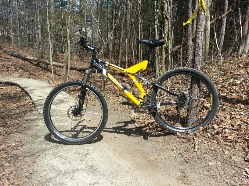 A mountain bike with a bright yellow frame is parked on a dirt trail surrounded by trees. The path winds through a forested area, with leaves scattered on the ground. A wooden boardwalk can be seen in the background, suggesting an outdoor recreational space. Georgia International Horse Park mountain bike trail.