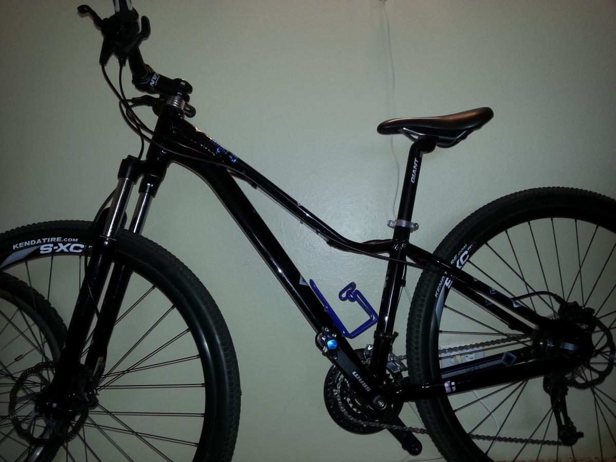 Giant 2012 Rainier 29er W: A black mountain bike with blue details, featuring front suspension, textured tires, and a sleek frame design, leaning against a light-colored wall.