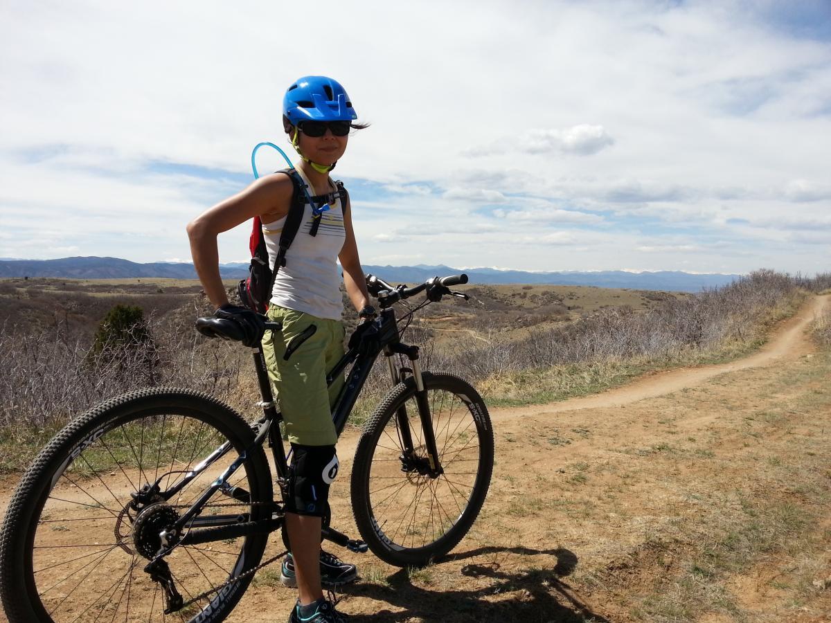 Giant 2012 Rainier 29er W: A person wearing a blue helmet and sunglasses stands beside a mountain bike on a dirt trail. They are dressed in a white tank top and green shorts, with protective knee pads. In the background, rolling hills and a clear blue sky can be seen, suggesting an outdoor biking adventure.