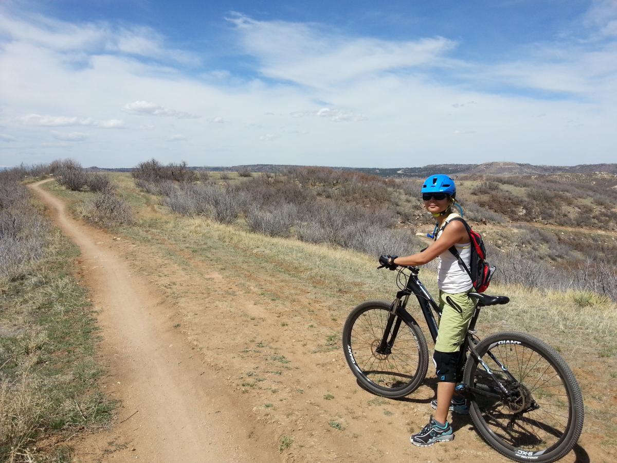Giant 2012 Rainier 29er W: A person in a blue helmet and athletic clothing stands beside a mountain bike on a dirt path, surrounded by a scenic landscape of hills and sparse vegetation under a partly cloudy sky.