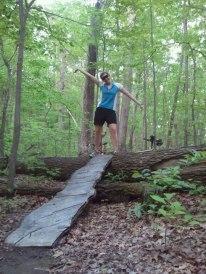 A person standing on a fallen log in a forest, with arms raised in a playful pose. A wooden plank is positioned on the log, leading across the forest floor covered in leaves and greenery. Sunlight filters through the dense tree canopy above. Kanawha State Forest Trails mountain bike trail.