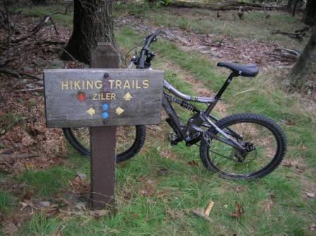 A wooden signpost marked "Hiking Trails Ziler" stands next to a mountain bike in a wooded area, with colorful trail markers indicating various routes. The ground is covered in grass and fallen leaves, and trees surround the scene. Cacapon State Park mountain bike trail.