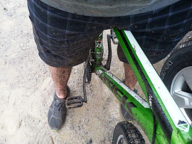 Trek Marlin: A person's legs, covered in mud, stand next to a green mountain bike. The individual is wearing shorts and gray shoes, with the bike pedal visible near their foot. The background shows a sandy surface and part of a vehicle tire.