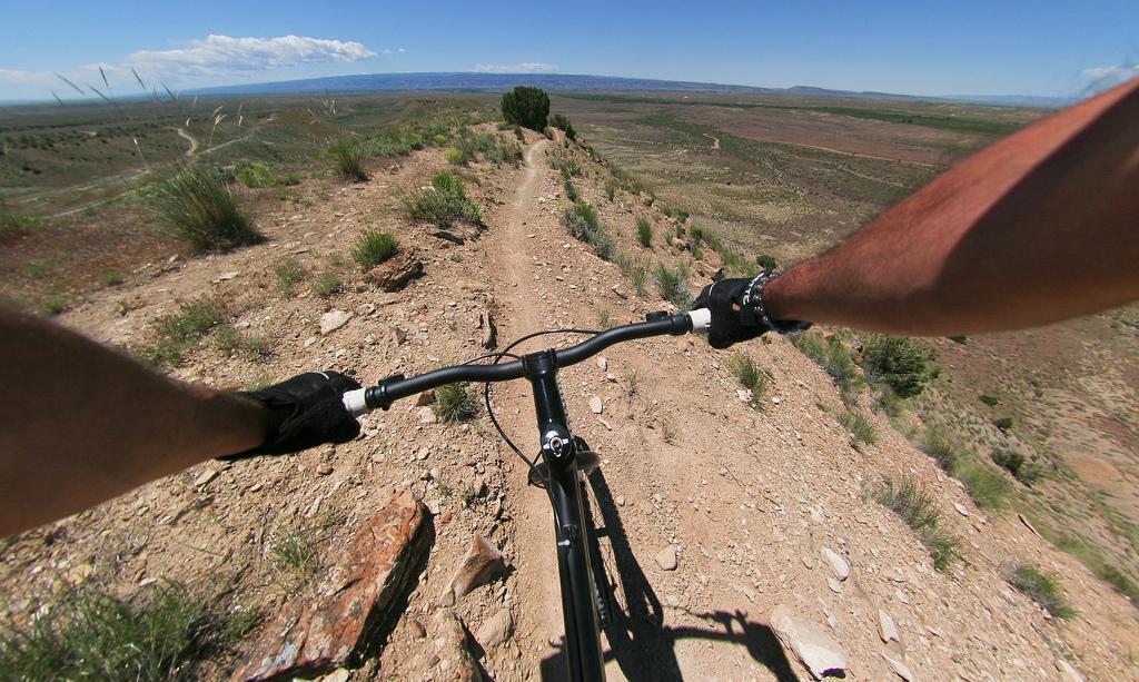 A view from the handlebars of a mountain bike riding down a rocky trail, with hands gripping the handlebars and an expansive landscape in the background, including hills and a blue sky. Zippety Do Dah mountain bike trail.