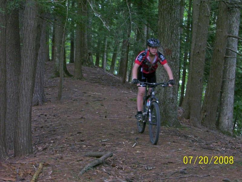 A person riding a mountain bike on a dirt path through a forest, surrounded by tall trees. The rider is wearing a helmet and a red and black cycling jersey, navigating the uneven terrain. North Bend State Park mountain bike trail.