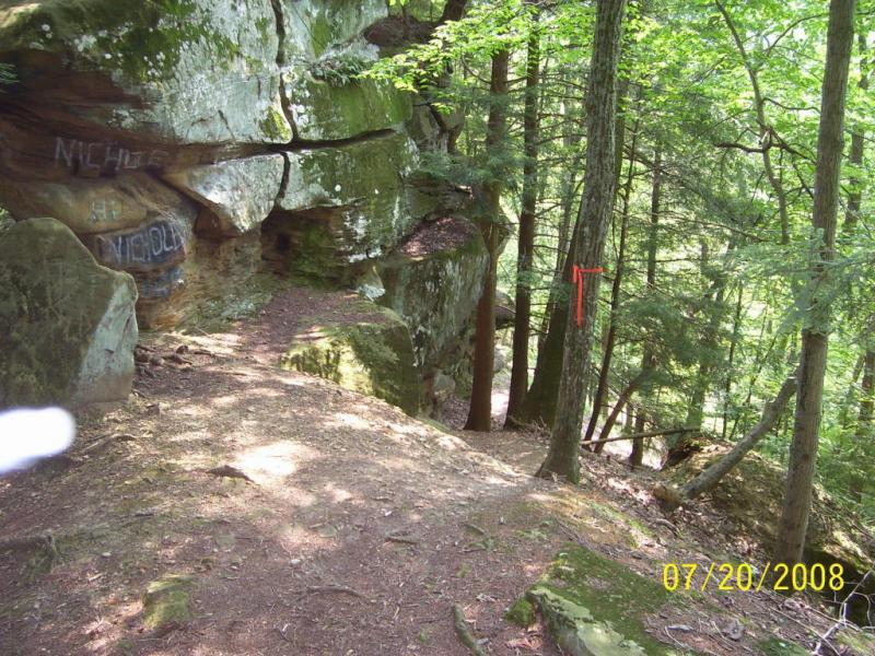 A rocky trail surrounded by lush green trees, with a large moss-covered boulder on the left side. Some graffiti is visible on the rock face, and a red ribbon is tied to a tree in the background. The scene conveys a sense of nature and outdoor exploration. North Bend State Park mountain bike trail.