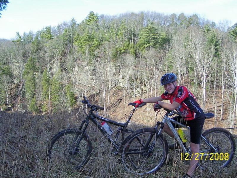 A young person in a red and black cycling jersey and helmet stands next to two mountain bikes on a grassy slope. They are smiling and leaning on one of the bikes, with a backdrop of a forested hillside featuring both evergreen and deciduous trees. The scene captures a sense of outdoor adventure and recreational biking. The date in the corner shows December 27, 2008. North Bend State Park mountain bike trail.