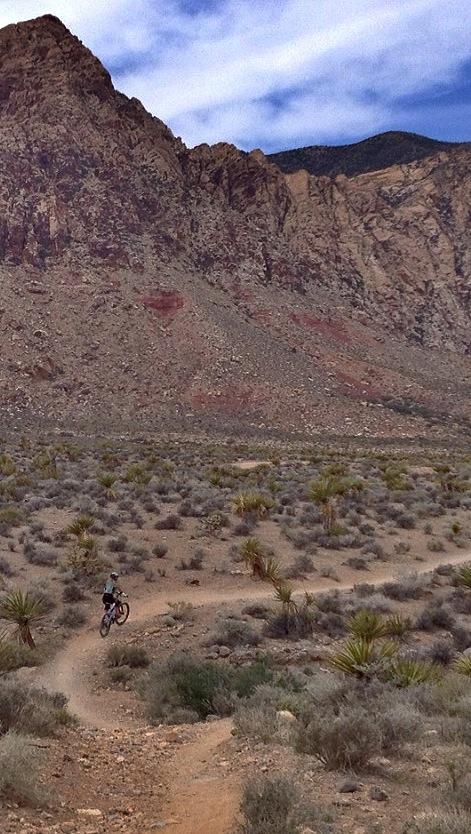 A person riding a mountain bike along a winding dirt trail in a desert landscape, with rocky cliffs and sparse vegetation in the background under a partly cloudy sky. Cottonwood Valley North mountain bike trail.