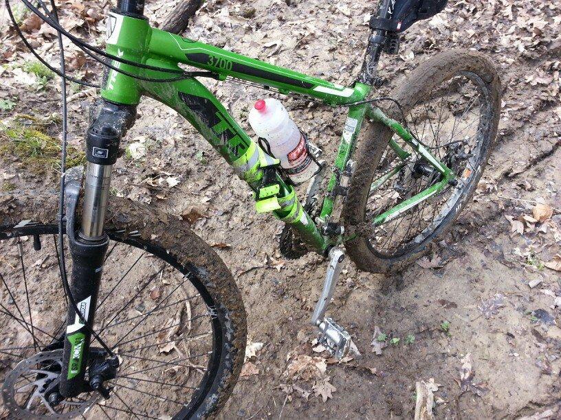 A close-up view of a green mountain bike with a muddy frame and tires, parked on a dirt trail surrounded by fallen leaves. A water bottle is attached to the bike frame, indicating preparation for outdoor riding. Wildlife Prairie Park mountain bike trail.