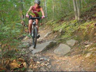 A person riding a mountain bike on a rocky trail in a forest, wearing a helmet and colorful clothing, with trees and greenery surrounding the path. Kanawha State Forest Trails mountain bike trail.