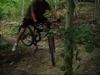 A person performing a bike jump in a wooded area, with a blurry motion effect. The scene shows tall trees and a dirt path, indicating an outdoor biking adventure. Kanawha State Forest Trails mountain bike trail.