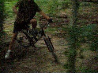 A person riding a bicycle through a wooded area, captured in motion. The background features trees and foliage, creating a natural setting. The cyclist is leaning to one side, indicating a dynamic movement on the trail. Kanawha State Forest Trails mountain bike trail.
