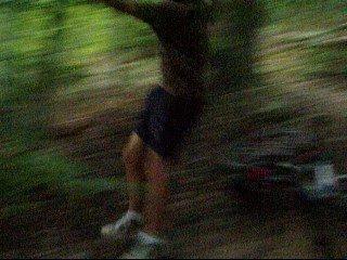 A person in a blurred motion, outdoors in a wooded area, wearing shorts and athletic shoes. The background features lush green foliage and ground cover, suggesting an active outdoor environment. Kanawha State Forest Trails mountain bike trail.