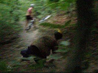 A blurred image depicting two individuals navigating a wooded trail. One person is seen in the background riding a bicycle, while the other is crouching on the path, surrounded by foliage. Kanawha State Forest Trails mountain bike trail.