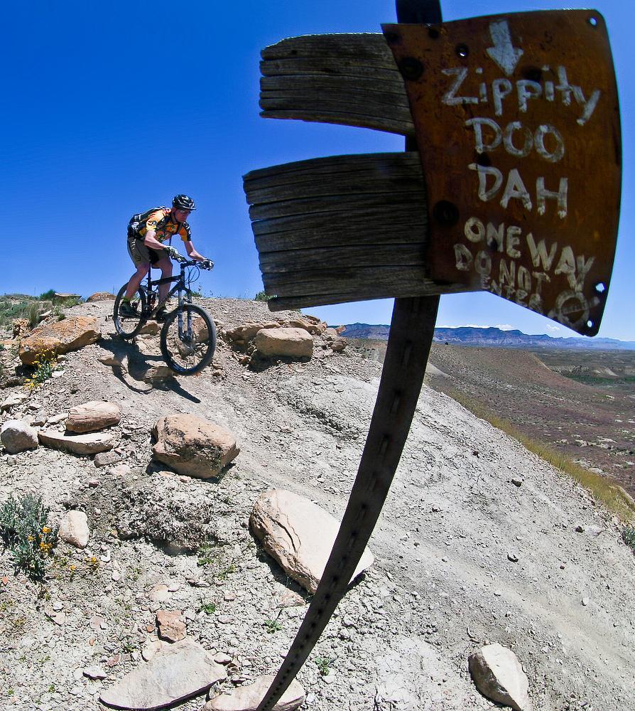 A mountain biker riding along a rocky trail, next to a wooden sign that reads "Zippity Doo Dah - One Way - Do Not Enter". The background features a clear blue sky and distant hills, highlighting a natural landscape. Zippety Do Dah mountain bike trail.