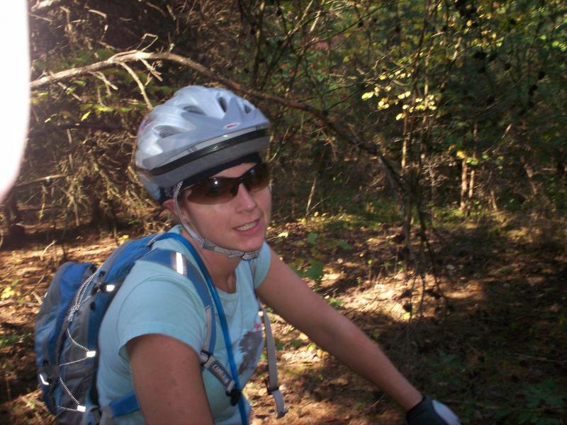 A woman wearing a bicycle helmet and sunglasses is sitting on a mountain bike in a wooded area. She has a backpack on her back and is looking towards the camera, smiling slightly. The surroundings include trees and foliage, indicating a natural outdoor setting. Panther Creek State Park mountain bike trail.