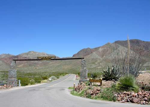 A dusty road leads under a stone archway with a wooden sign that reads "Welcome to Water Canyon." The surrounding landscape features rolling hills and mountains, with sparse vegetation including cacti and shrubs under a clear blue sky. Franklin Mountain State Park mountain bike trail.