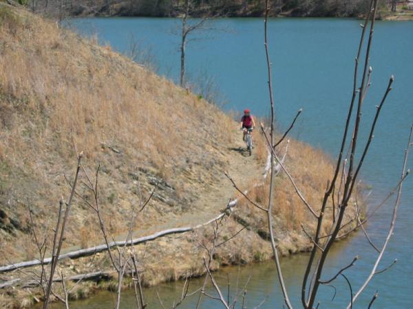 A cyclist riding along a narrow, grassy trail near the edge of a calm blue lake, surrounded by sparse trees and dry foliage. Douthat State Park mountain bike trail.